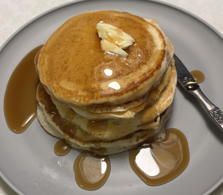 A stack of fluffy pancakes, perfect for budget meals, is topped with melting butter and drizzled with syrup on a gray plate, with a knife resting on the side and syrup pooled around the pancakes.