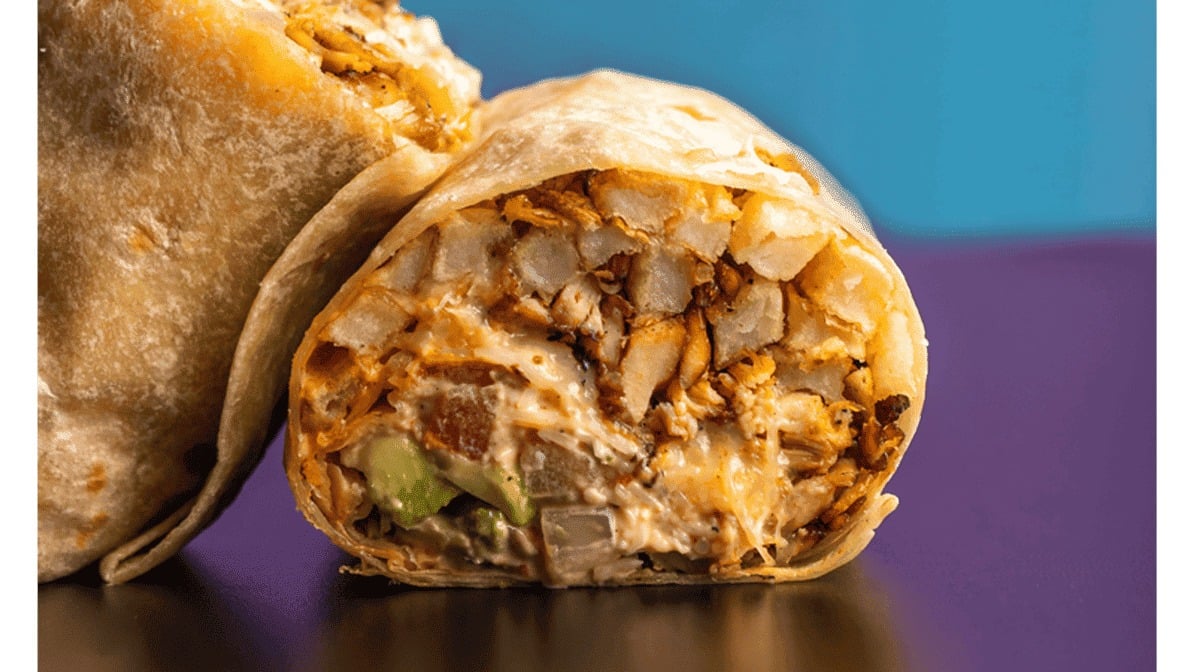 A close-up of a burrito cut in half reveals layers of diced potatoes, shredded chicken, melted cheese, creamy sauce, and avocado in a flour tortilla—a feast reminiscent of top breakfast burrito spots—set against a bright blue and purple background.