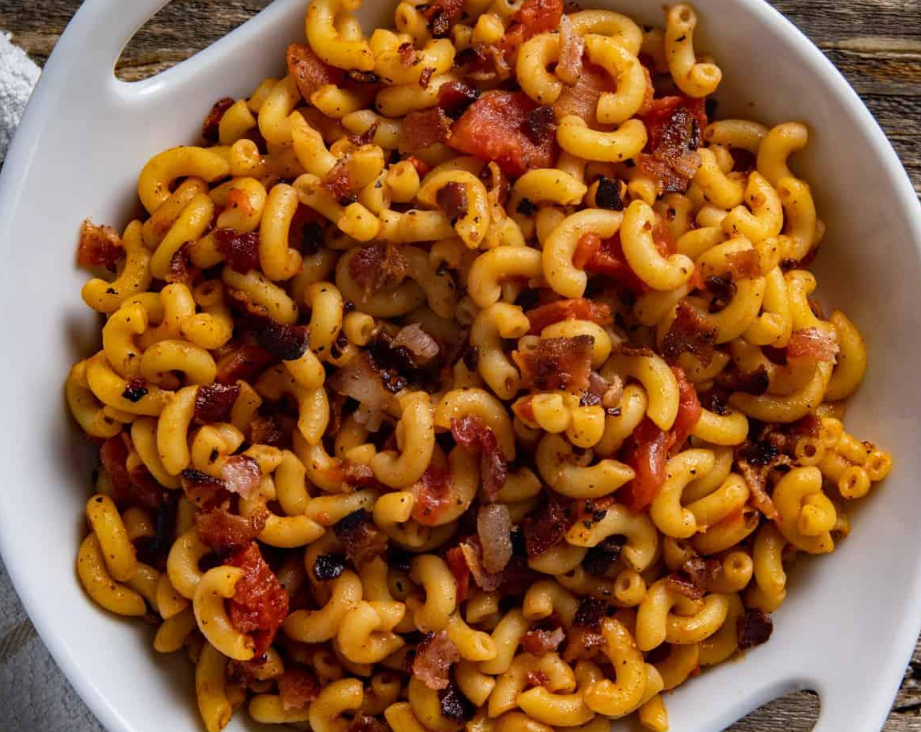 A white bowl filled with cooked elbow macaroni mixed with crispy bacon pieces and diced tomatoes, placed on a wooden surface—an easy recipe perfect for delicious budget meals.