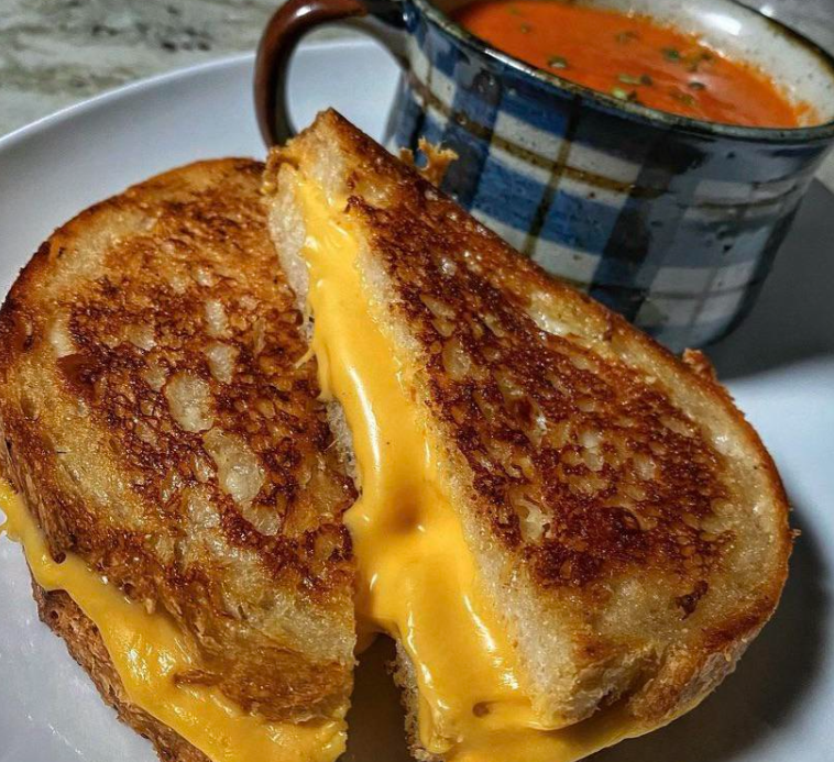 A golden brown grilled cheese sandwich with melted cheese is sliced in half and served on a white plate, next to a mug of creamy tomato soup—classic comfort food that's perfect for budget meals.