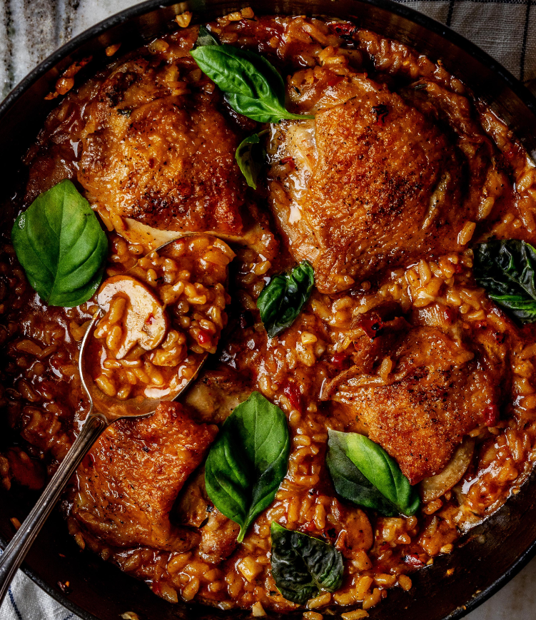 A skillet filled with golden-brown chicken thighs, creamy rice in a rich sauce, and garnished with fresh basil—this delicious budget meal is ready to serve, with a spoon resting in the rice.