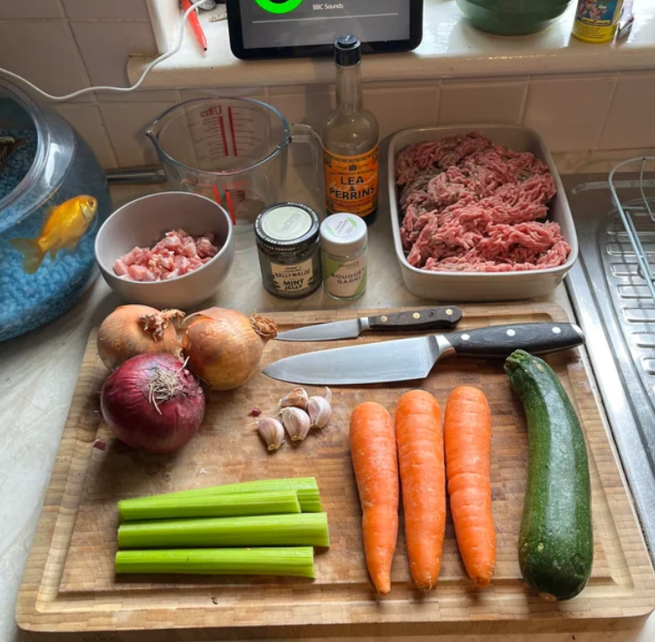 A chopping board with knives, red and yellow onions, garlic, celery, carrots, and zucchini—perfect ingredients for budget meals. Nearby are bowls of minced meat, diced bacon, a measuring jug, jars, Worcestershire sauce—and a goldfish in a tank.