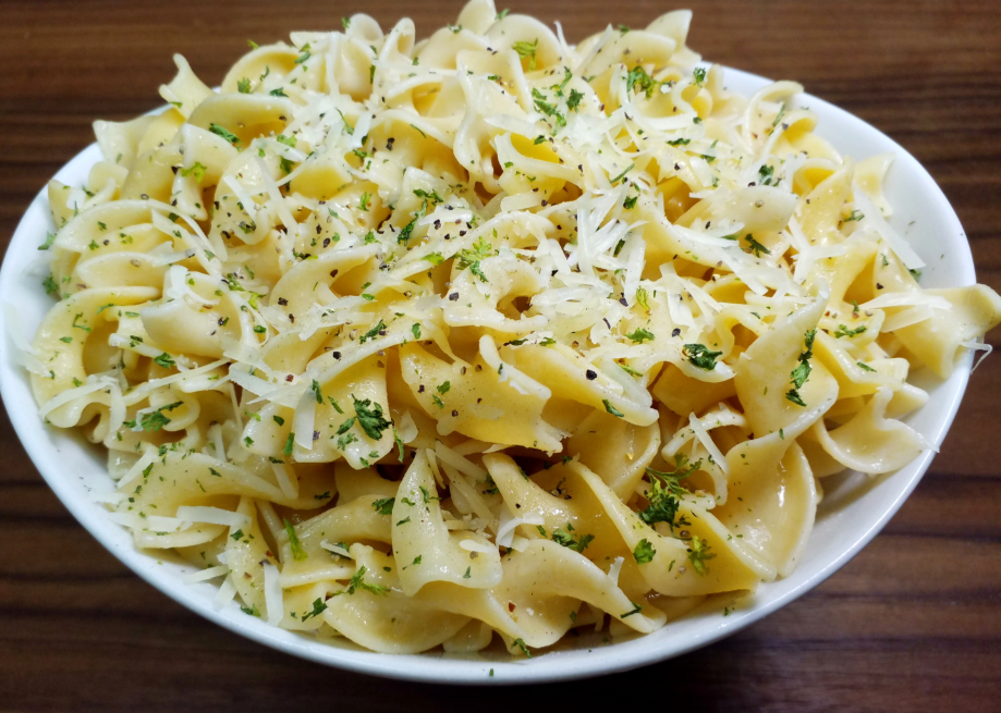 A white bowl filled with cooked egg noodles, topped with grated cheese and chopped herbs, sits on a wooden surface—an inviting example of delicious budget meals.