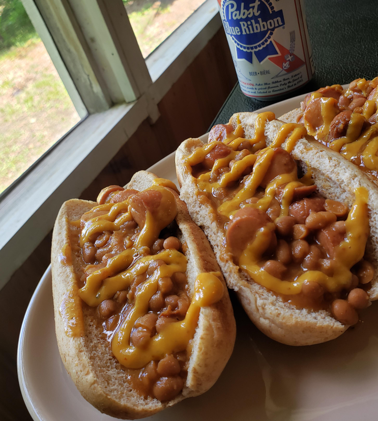 Two hot dogs topped with baked beans, sliced sausage, and melted cheddar cheese sit on a plate by a window—an easy budget meal. A can of Pabst Blue Ribbon beer is in the background.