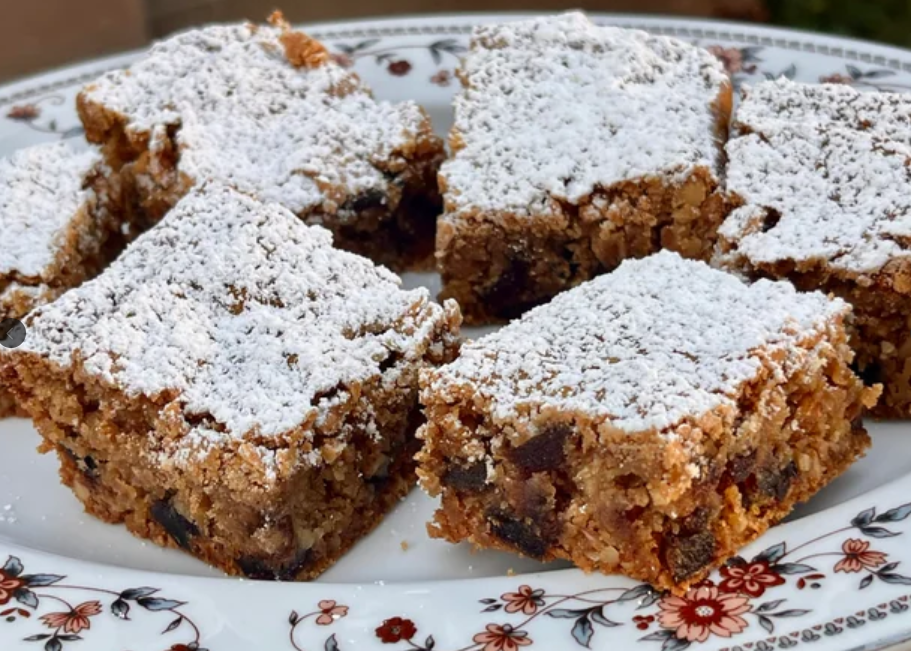 Six square pieces of date nut bars, perfect for budget meals, are topped with powdered sugar and arranged on a floral-patterned plate. The bars appear moist with visible bits of dates and nuts.