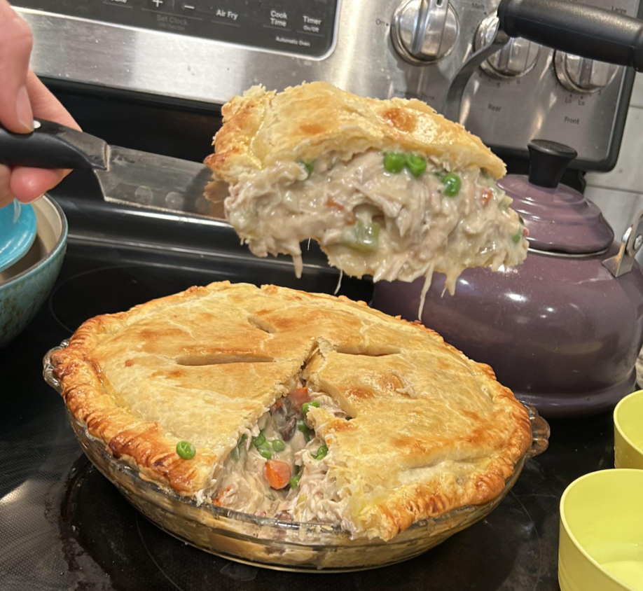 A hand lifts a slice of creamy chicken pot pie with vegetables from a baking dish. The golden, flaky crust is partially cut, revealing a filling of chicken, peas, and carrots—an ideal choice for comforting budget meals. Kitchen appliances are visible in the background.