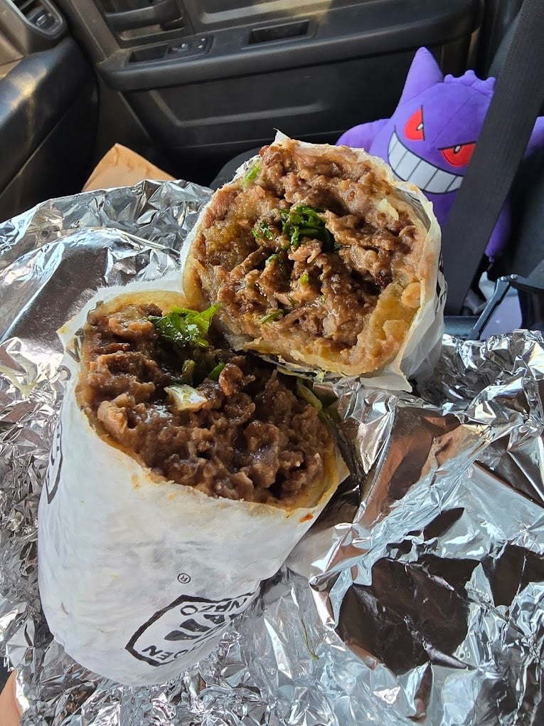 A foil-wrapped burrito cut in half, filled with meat, beans, and greens, sits on a car seat—ready for a morning bite. In the background, a plush Gengar toy is buckled in, perhaps dreaming of the best breakfast burrito spots.