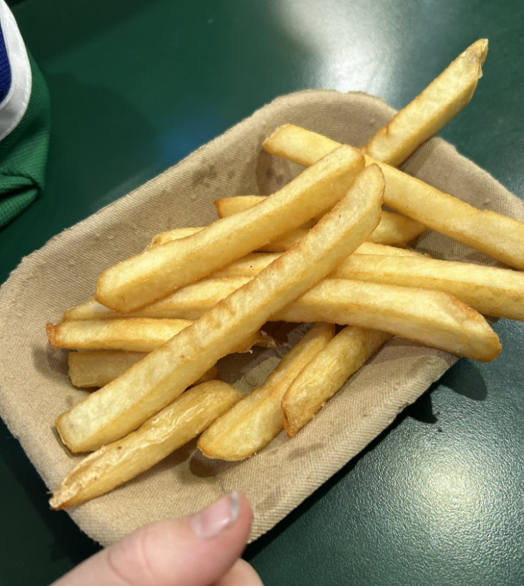A small rectangular basket lined with brown paper holds a serving of golden, crispy French fries. A thumb and fingers are visible at the bottom of the image, holding the basket against a green tabletop.