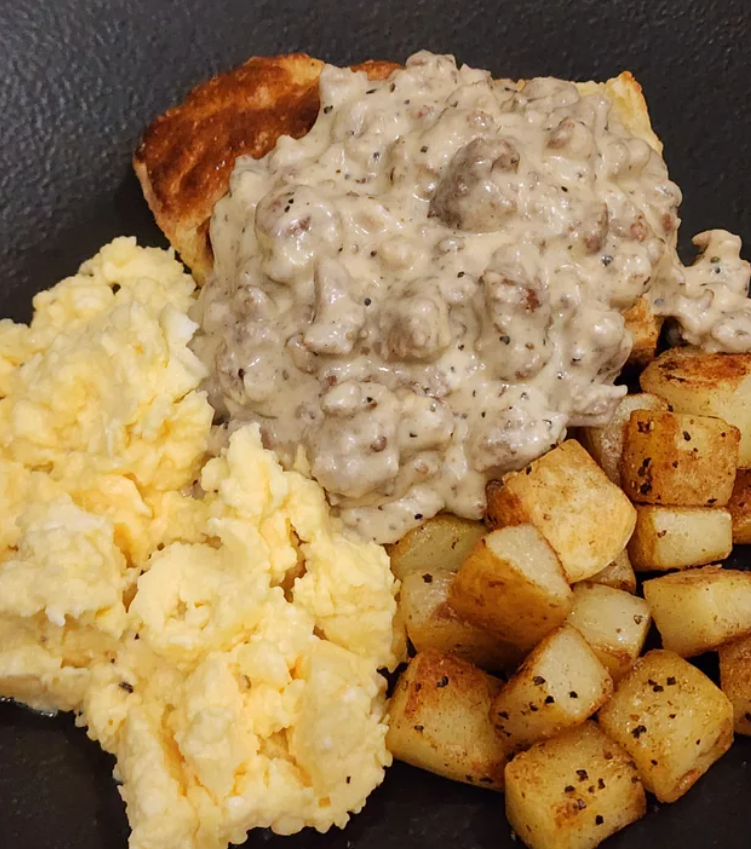 A hearty breakfast plate with scrambled eggs, golden-brown diced potatoes, and a biscuit topped with creamy sausage gravy—perfect for those seeking delicious budget meals on a dark background.