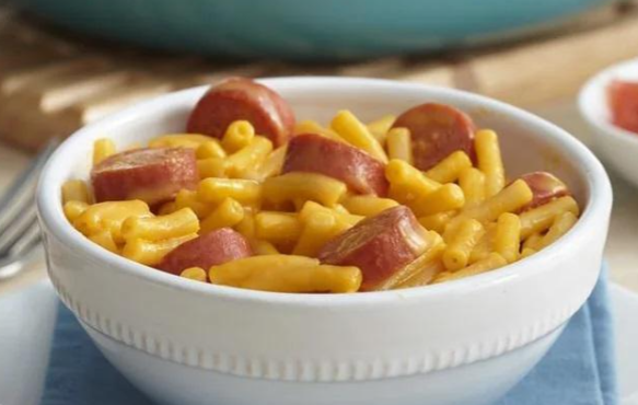 A white bowl filled with macaroni and cheese, topped with sliced pieces of sausage or hot dog—a classic budget meal. The bowl sits on a blue napkin, with a blurry plate visible in the background.