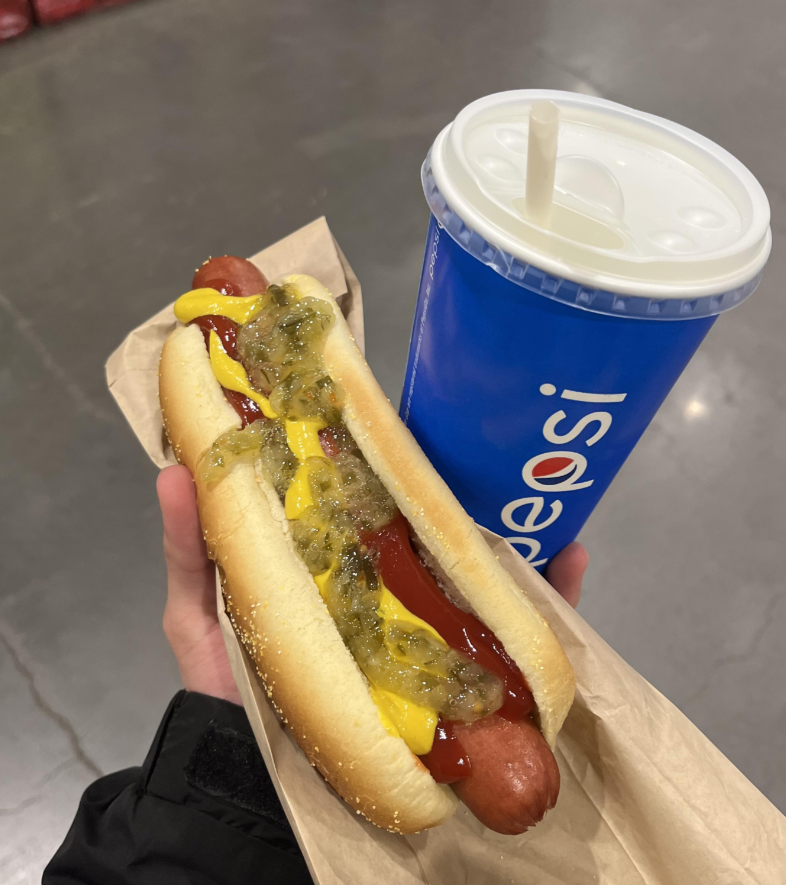 A hand holds a hot dog topped with ketchup, mustard, and relish in a paper sleeve, next to a blue Pepsi cup with a straw—classic staples of fast-food value meals. The background reveals a gray indoor floor.