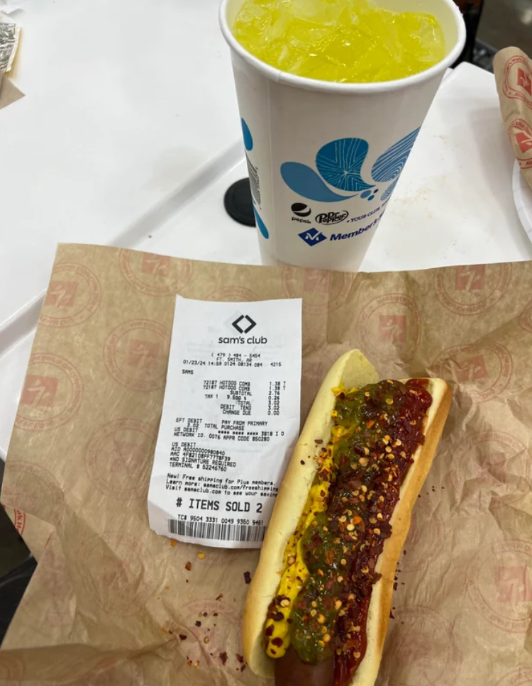 A hot dog with mustard, relish, and ketchup sits on brown paper next to a Sam’s Club receipt and a large cup of bright yellow-green soda—classic elements of fast-food value meals enjoyed on the go.