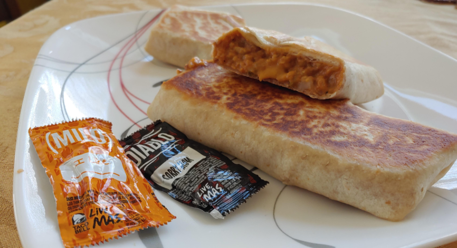 A plate holds two grilled burritos, one cut open to reveal a cheesy, saucy filling—perfect for fans of fast-food value meals. Next to them are Taco Bell sauce packets labeled "Mild" and "Diablo.