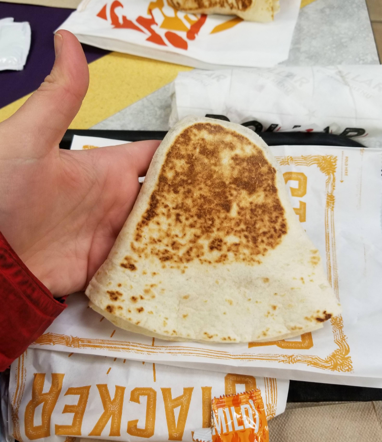 A hand holding a folded Taco Bell quesadilla or Crunchwrap Supreme, with Taco Bell branded wrappers and a mild sauce packet on a fast-food value meals tray in the background.