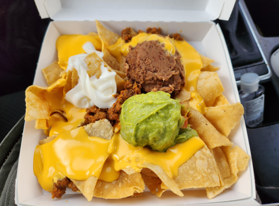 A takeout container filled with nachos topped with melted cheese sauce, ground beef, refried beans, guacamole, and a dollop of sour cream sits inside a car—capturing the crave-worthy comfort of fast-food value meals on the go.