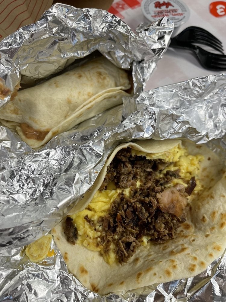 Two breakfast burritos wrapped in foil, one opened to reveal scrambled eggs and crumbled sausage in a flour tortilla. Plastic forks and napkins are visible—just what you’d expect from top breakfast burrito spots.