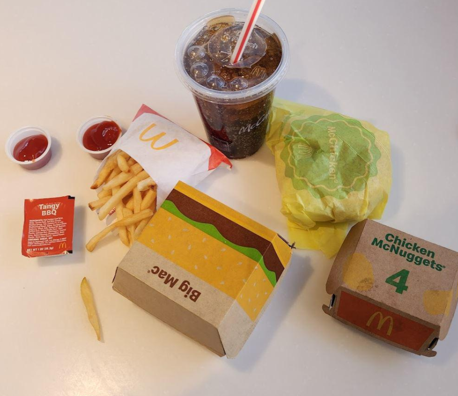 A classic fast-food value meal featuring a drink, fries, ketchup, Tangy BBQ sauce, a wrapped burger, and a box of 4 Chicken McNuggets, all neatly arranged on a light-colored table.