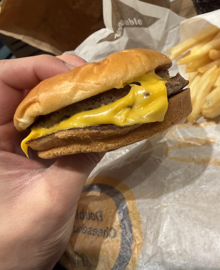 A hand holding a cheeseburger with melted cheese and a beef patty in a soft bun, part of one of the classic fast-food value meals. French fries and branded fast food packaging are visible in the background.