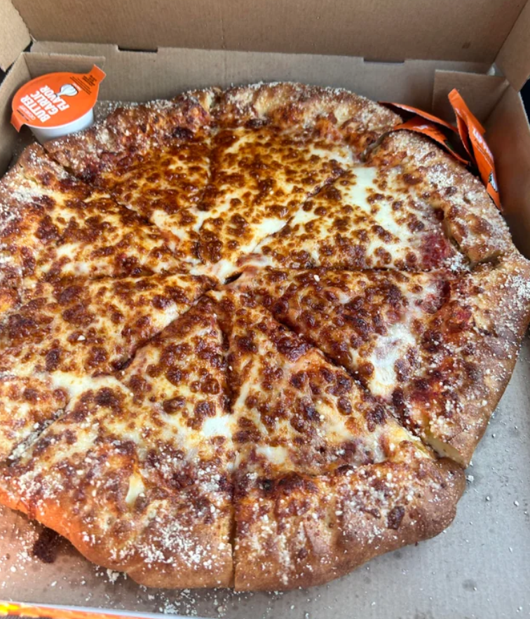 A close-up of a cheesy pizza with a thick, seasoned crust, cut into eight slices and served in an open cardboard pizza box—just like the ones found in popular fast-food value meals. Dipping sauce containers are visible in the corners of the box.