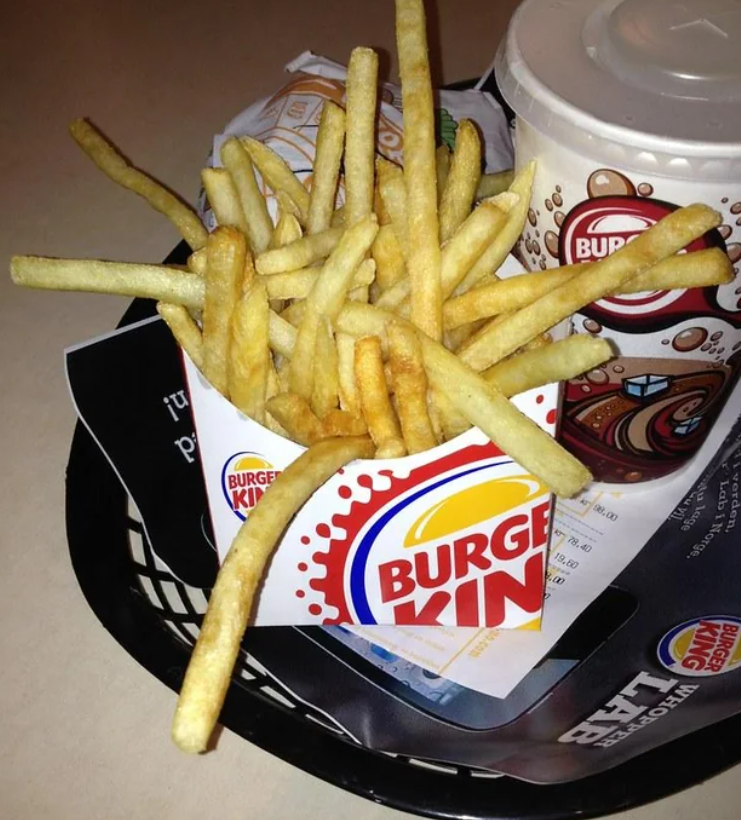 A serving of Burger King French fries in a branded container sits next to a Burger King soft drink cup on a tray, showcasing the appeal of fast-food value meals with food wrappers and a receipt underneath.