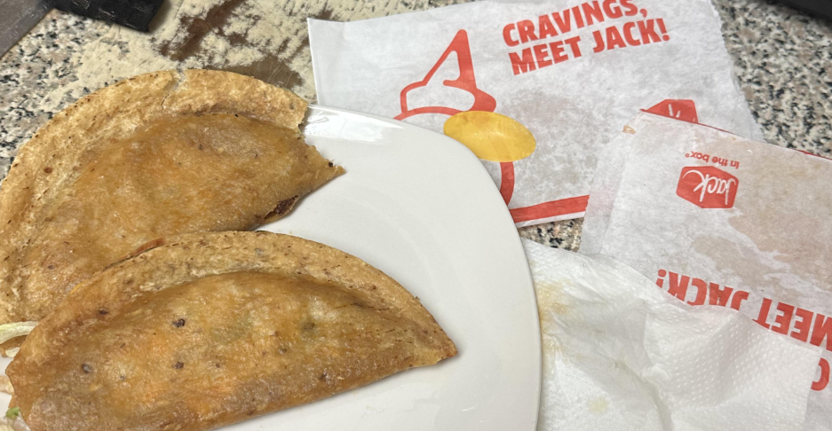 Two crispy taco shells filled with meat on a white plate, next to Jack in the Box branded wrappers and a napkin, showcase classic fast-food value meals on a speckled countertop.