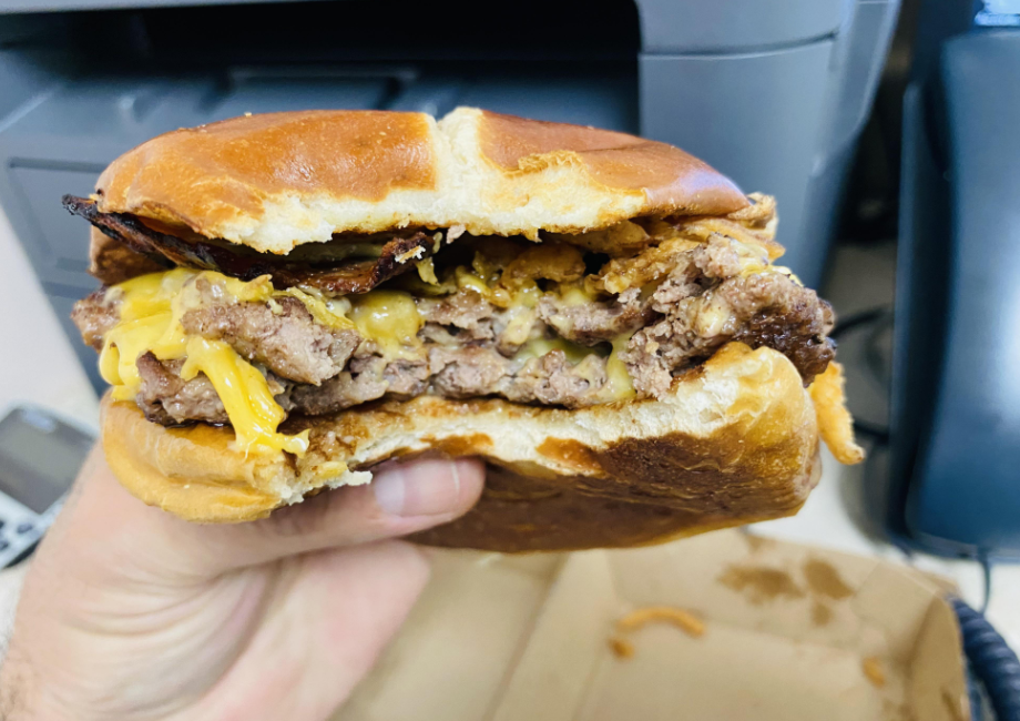 A hand holds a cheeseburger with two beef patties, melted cheddar cheese, crispy onions, and a toasted bun—an irresistible pick among fast-food value meals. Office equipment and a takeout box are visible in the blurred background.