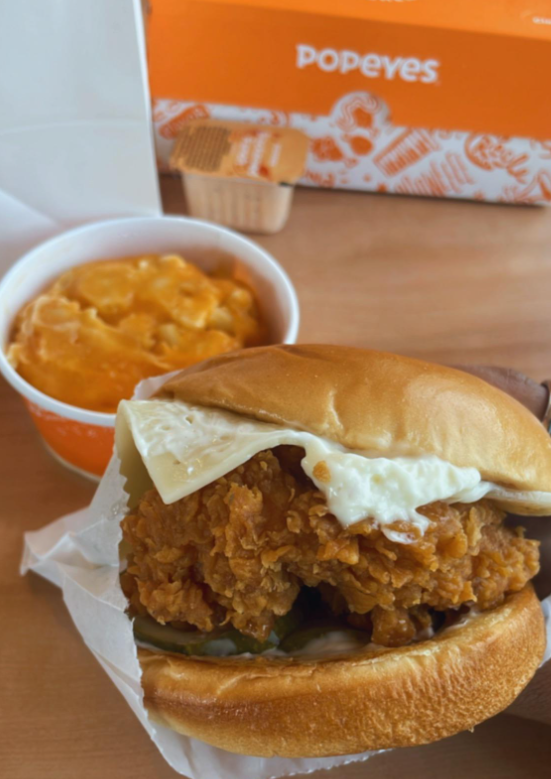 A crispy fried chicken sandwich with a slice of cheese sits on a wrapper beside creamy macaroni and cheese—classic fast-food value meals. In the background, a Popeyes box and sauce packet rest on a wooden surface.