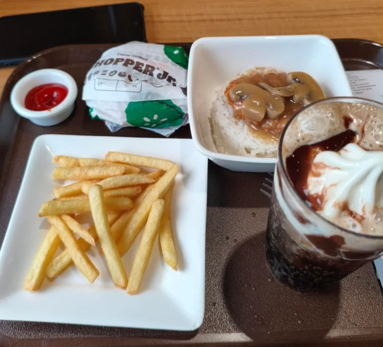 A fast-food value meal featuring fries, ketchup, a wrapped burger, mashed potatoes with gravy and mushrooms, and a glass of ice cream float, all served on a brown tray.