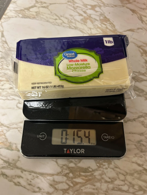 A block of Great Value whole milk low-moisture mozzarella cheese sits on a digital kitchen scale, displaying a weight of 15.4 ounces on a marble countertop.