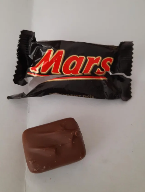A small, unwrapped Mars chocolate bar sits on a white surface next to its black and red Mars wrapper.
