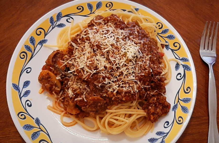 A plate of spaghetti topped with meat sauce and grated cheese, reminiscent of meals middle-class moms cooked, served on a decorative yellow and blue patterned plate next to a fork on a wooden table.