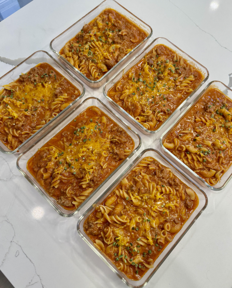 Six glass meal prep containers filled with cooked rotini pasta, ground meat, and tomato sauce, topped with melted cheese—delicious meals middle-class moms cooked and arranged neatly on a white countertop.