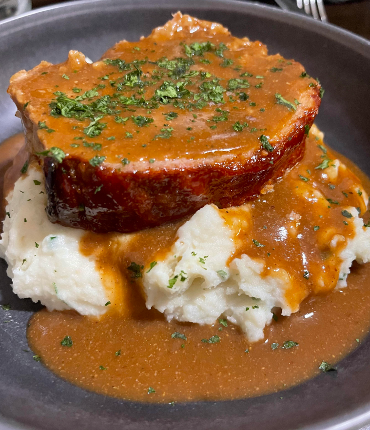 A thick slice of meatloaf topped with brown gravy and chopped herbs, served on a generous portion of mashed potatoes with extra gravy—a classic dish reminiscent of meals middle-class moms cooked at home.