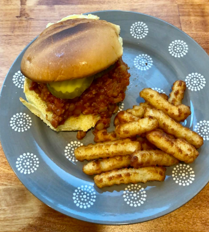 A gray plate with a sloppy joe sandwich—one of those classic meals middle-class moms cooked—topped with pickles and served with crispy crinkle-cut fries, all set on a wooden table.