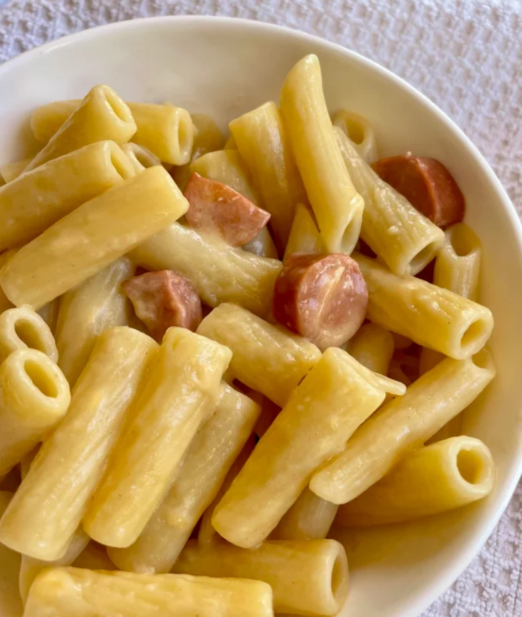 A close-up of a bowl filled with rigatoni pasta and sliced sausage pieces, coated in a creamy yellow sauce, evokes comforting meals middle-class moms cooked, placed on a textured white surface.
