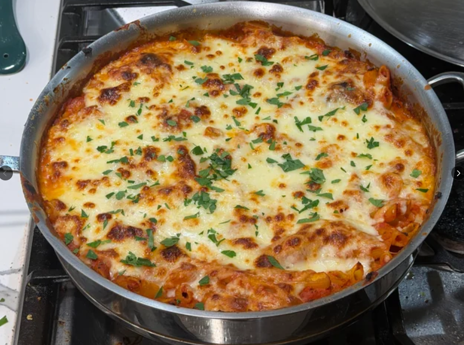 A skillet filled with baked pasta topped with melted, golden-brown cheese and sprinkled with chopped fresh parsley, reminiscent of the comforting meals middle-class moms cooked, sits invitingly on the stove.