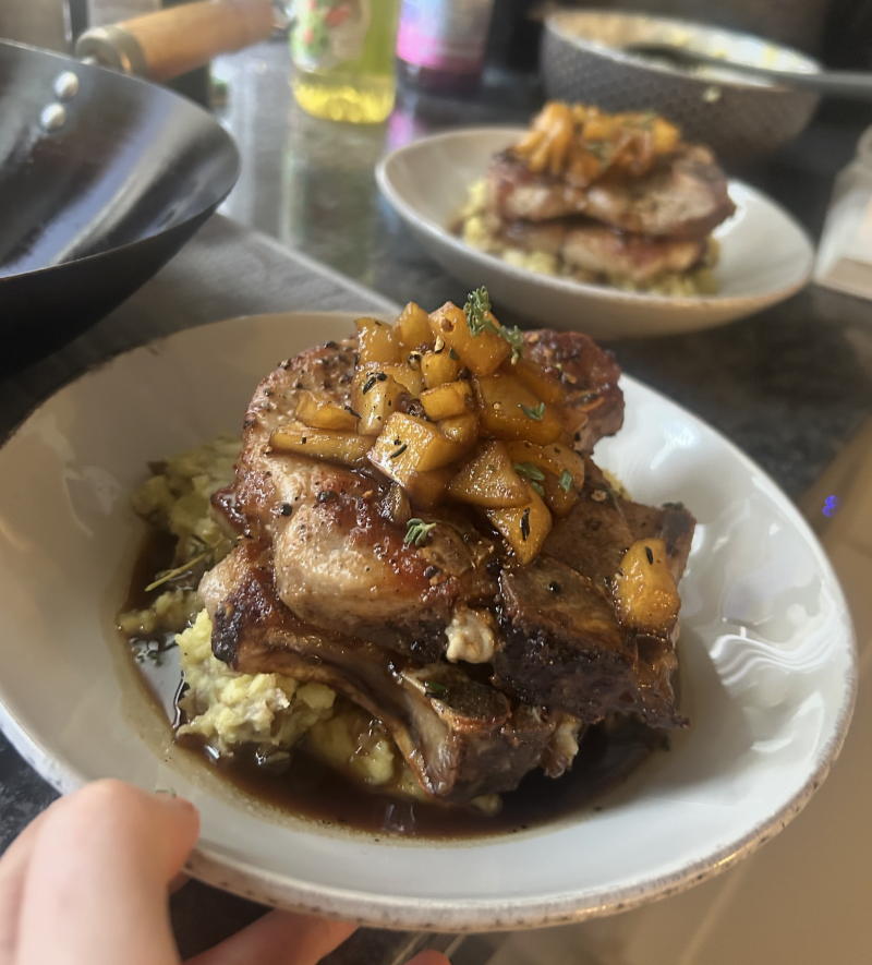 A plate of grilled pork chops topped with diced apples and herbs, served over mashed potatoes—a classic example of the hearty meals middle-class moms cooked—sits on a kitchen counter among various cooking utensils, with another plate blurred in the background.
