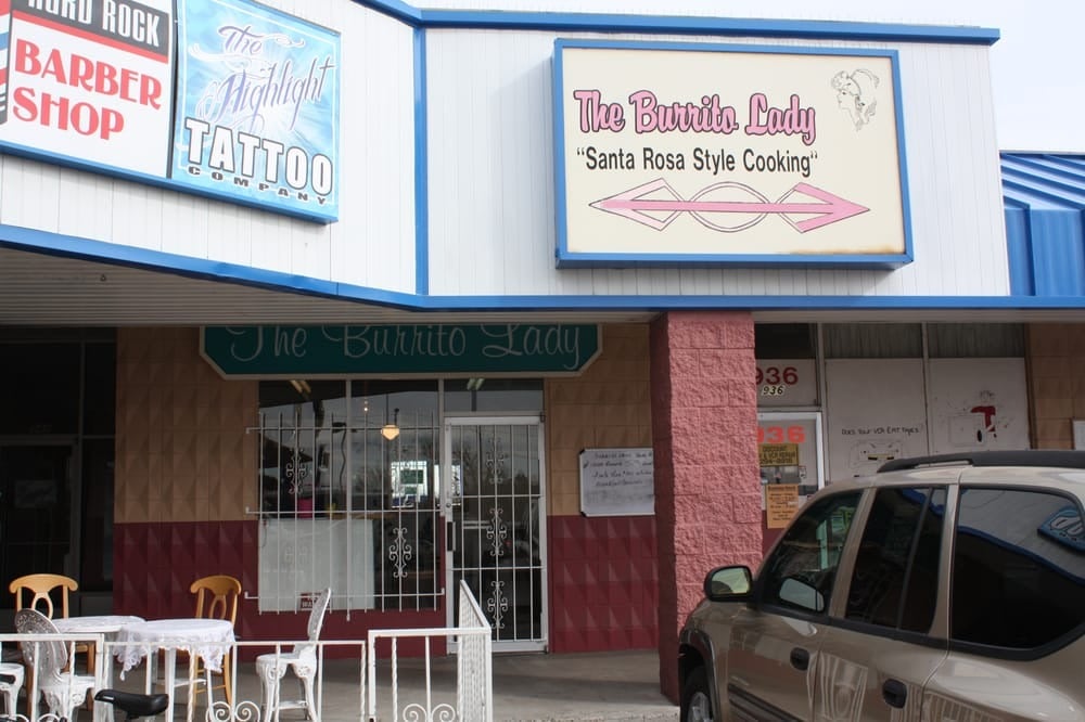Storefront of "The Burrito Lady" restaurant with a sign reading "Santa Rosa Style Cooking"; outdoor tables and chairs are visible, with a vehicle parked in front—one of the local breakfast burrito spots. Adjacent signs advertise a barber shop and tattoo company.