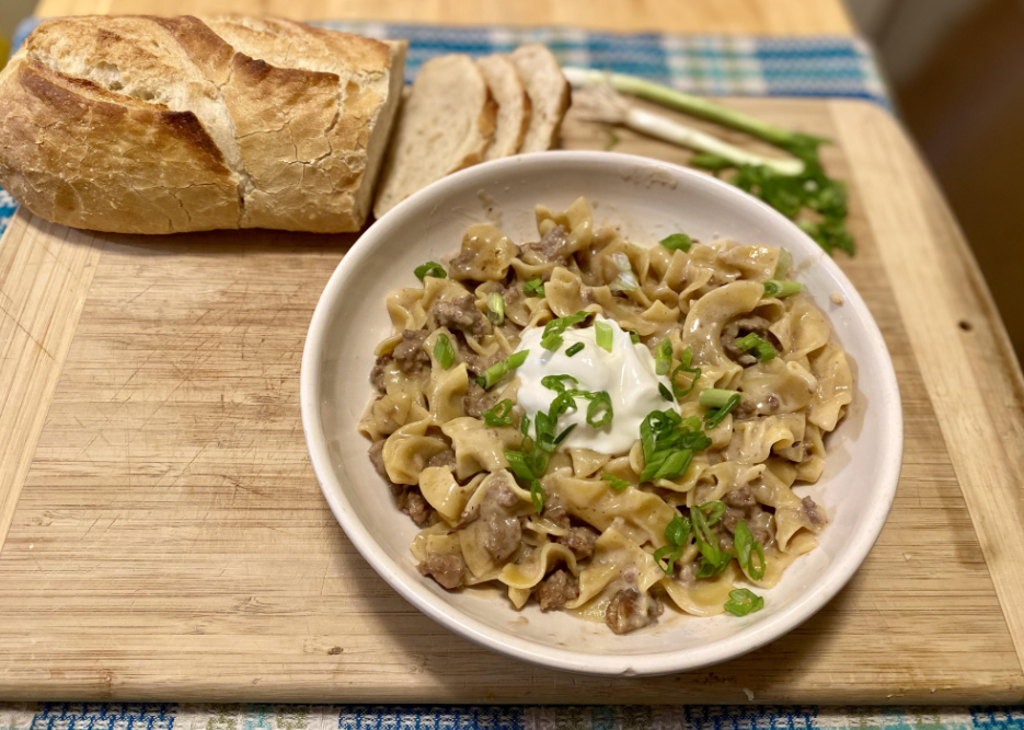 A bowl of beef stroganoff with egg noodles, topped with sour cream and chopped green onions, sits on a wooden board next to sliced baguette—a classic example of meals middle-class moms cooked at home.