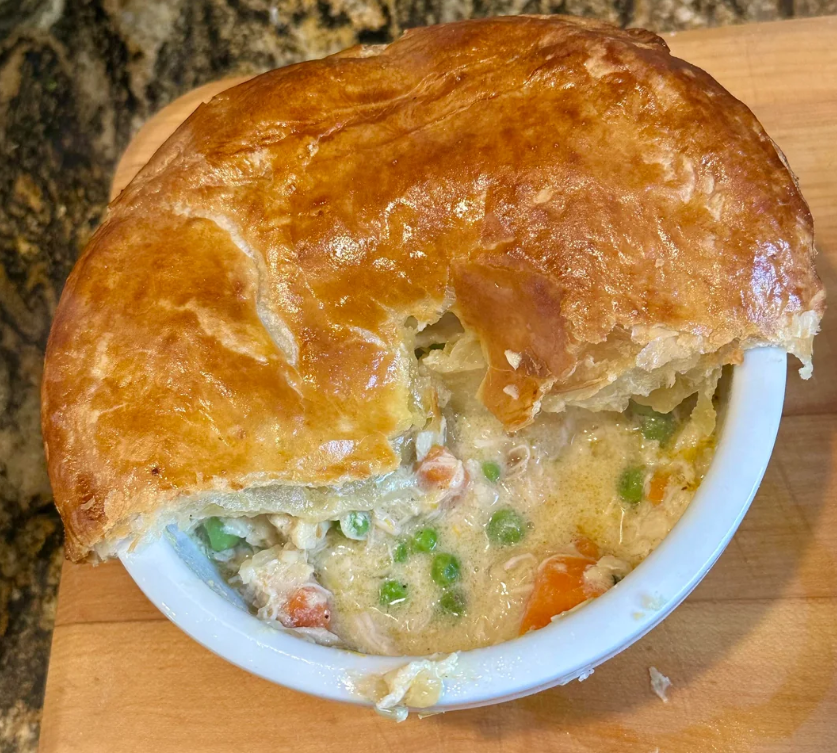 A golden-brown puff pastry-topped chicken pot pie, reminiscent of the comforting meals middle-class moms cooked, with a portion missing to reveal creamy chicken, peas, carrots, and vegetables in a white ramekin on a wooden surface.