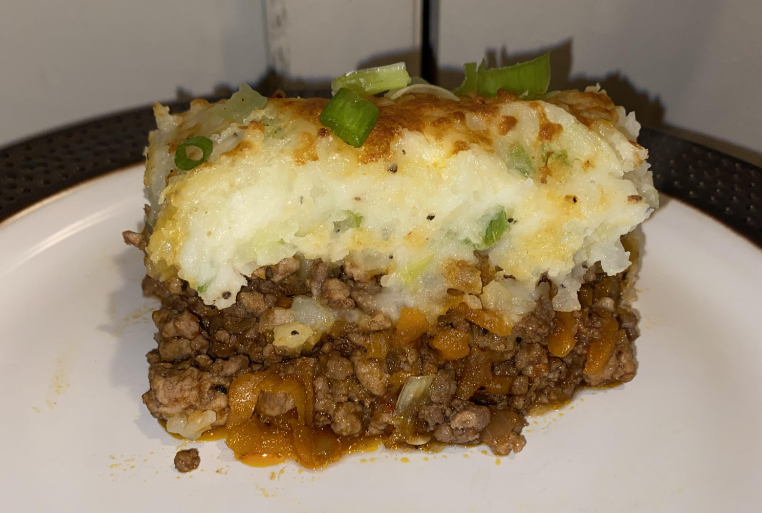 A slice of shepherd’s pie on a white plate, showing layers of ground meat, carrots, and onions—classic comfort food topped with mashed potatoes, melted cheese, and green onions—just like the hearty meals middle-class moms cooked.