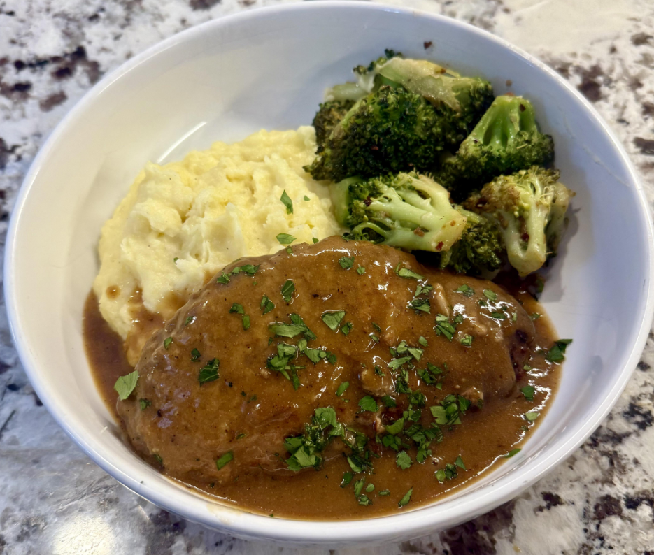 A bowl with mashed potatoes, broccoli florets, and a piece of meatloaf topped with brown gravy and chopped herbs—a classic example of the comforting meals middle-class moms cooked, set on a granite countertop.