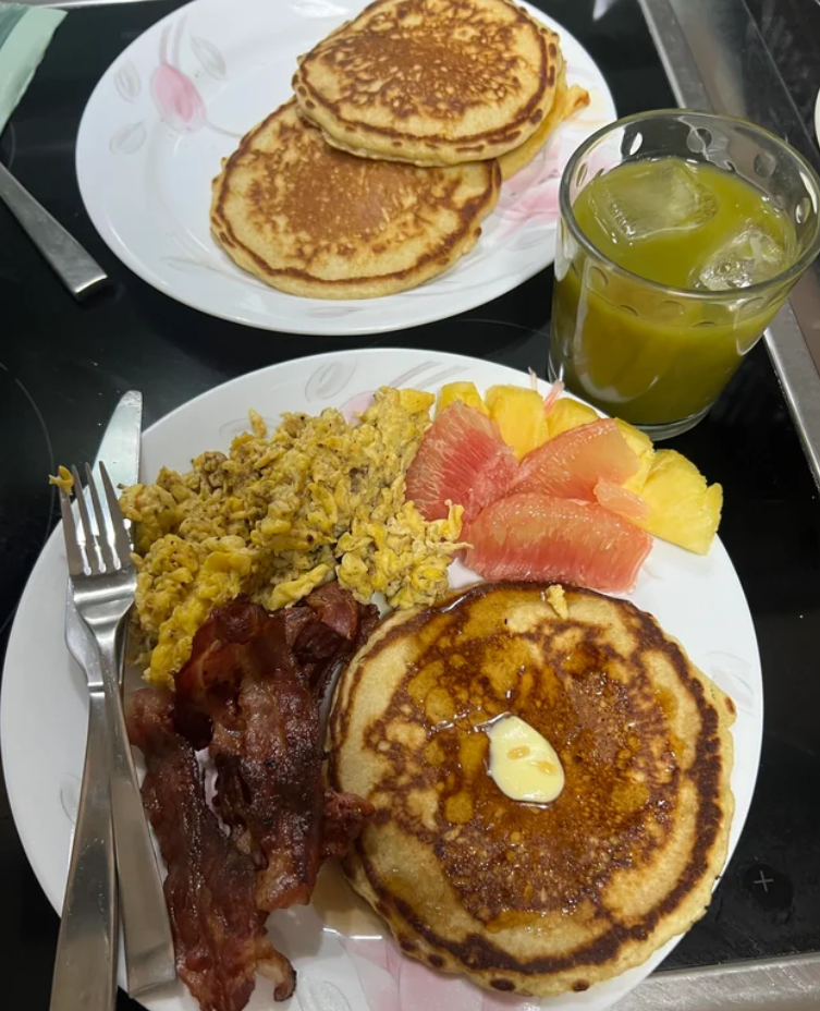 A plate with pancakes topped with butter, scrambled eggs, crispy bacon, grapefruit, and pineapple slices—reminiscent of the hearty meals middle-class moms cooked. Behind it sits more pancakes and a glass of iced green juice. Cutlery rests on the front plate.