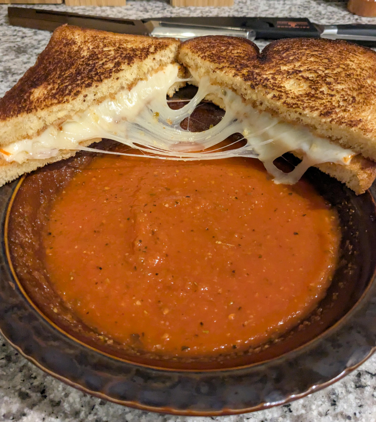 A grilled cheese sandwich with gooey, melted cheese—like the classic meals middle-class moms cooked—is split in half and resting on the edge of a bowl filled with creamy tomato soup. The cheese stretches between the halves over the soup.