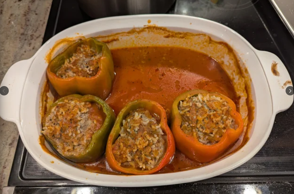 Four stuffed bell peppers, two green and two orange, filled with a mixture of rice and ground meat, sit in a white baking dish surrounded by tomato sauce—a classic example of the hearty meals middle-class moms cooked.
