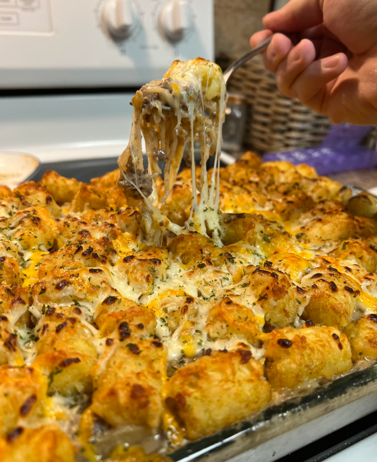 A hand lifts a serving of cheesy tater tot casserole with a fork, showing melted cheese stretching from a baking dish filled with golden tater tots and toppings—a classic comfort food often found among meals middle-class moms cooked.