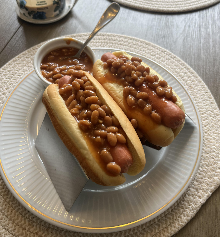 Two hot dogs in buns topped with baked beans sit in a metal holder on a white plate, recalling the comforting meals middle-class moms cooked, with a bowl of extra baked beans and a spoon beside them on a woven placemat.