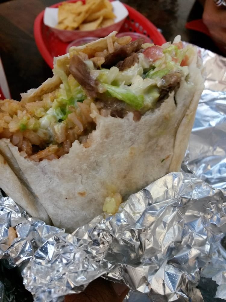 A close-up of a partially unwrapped burrito from one of the top breakfast burrito spots, filled with rice, lettuce, meat, and more, rests on crumpled aluminum foil. In the background, there’s a red basket with tortilla chips.