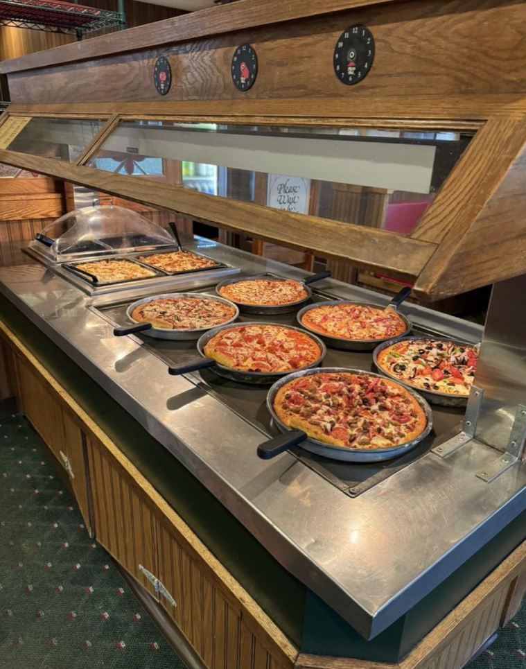 A buffet counter displays several varieties of pizza from popular pizza chains in round pans, including cheese, pepperoni, and pizzas with mixed toppings, under a wooden and glass sneeze guard.