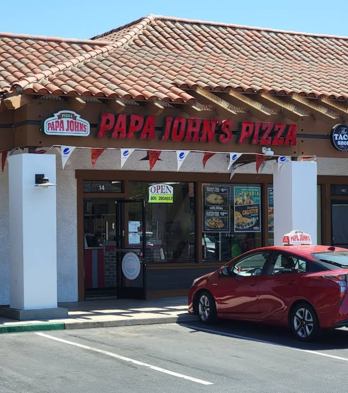 A Papa John’s Pizza storefront, part of popular pizza chains, features a red car parked in front. The shop has a tiled roof, an open sign on the door, pizza ads in the windows, and red pennant flags above the entrance.